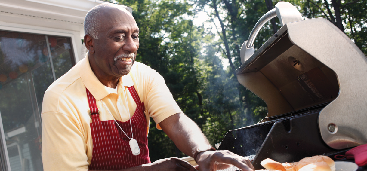 An older black man wearing the Auto Alert fall detection button is using the barbecue outside.