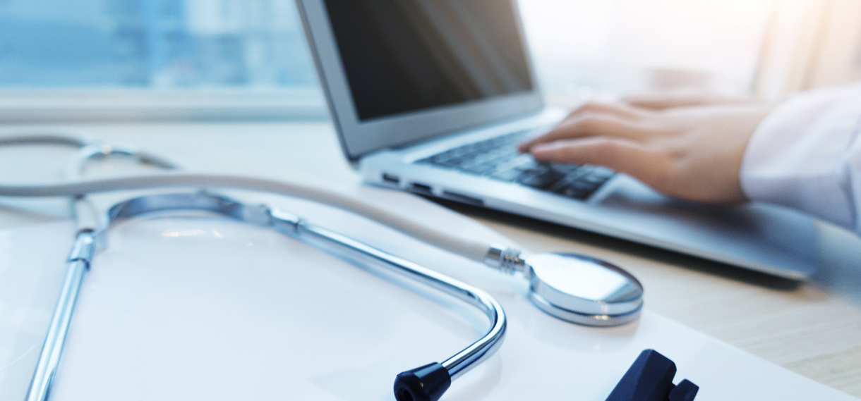 Someone typing on a computer with a stethoscope next to them on the desk.