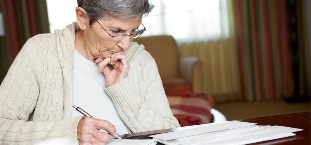 Older woman sitting at the table paying bills with a worried look on her face.