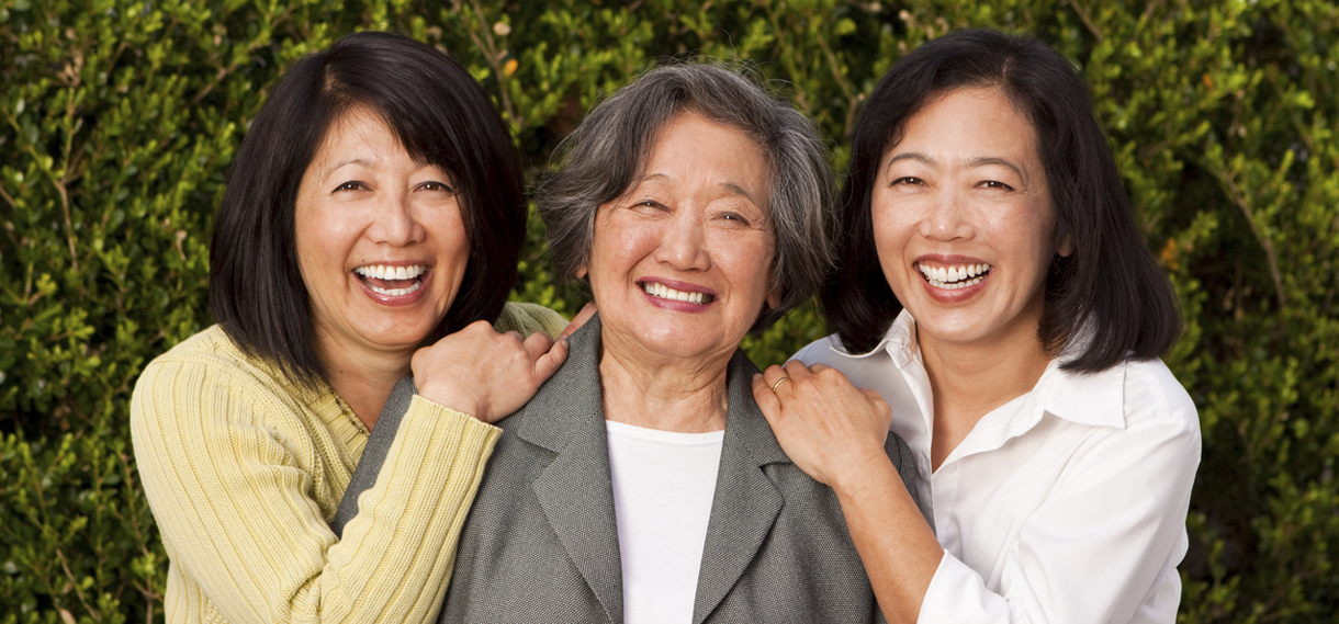 Asian mother and her two adult daughters smiling in front some evergreen trees.