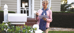 An older woman wearing a Go Safe mobile button is standing next to her mailbox.