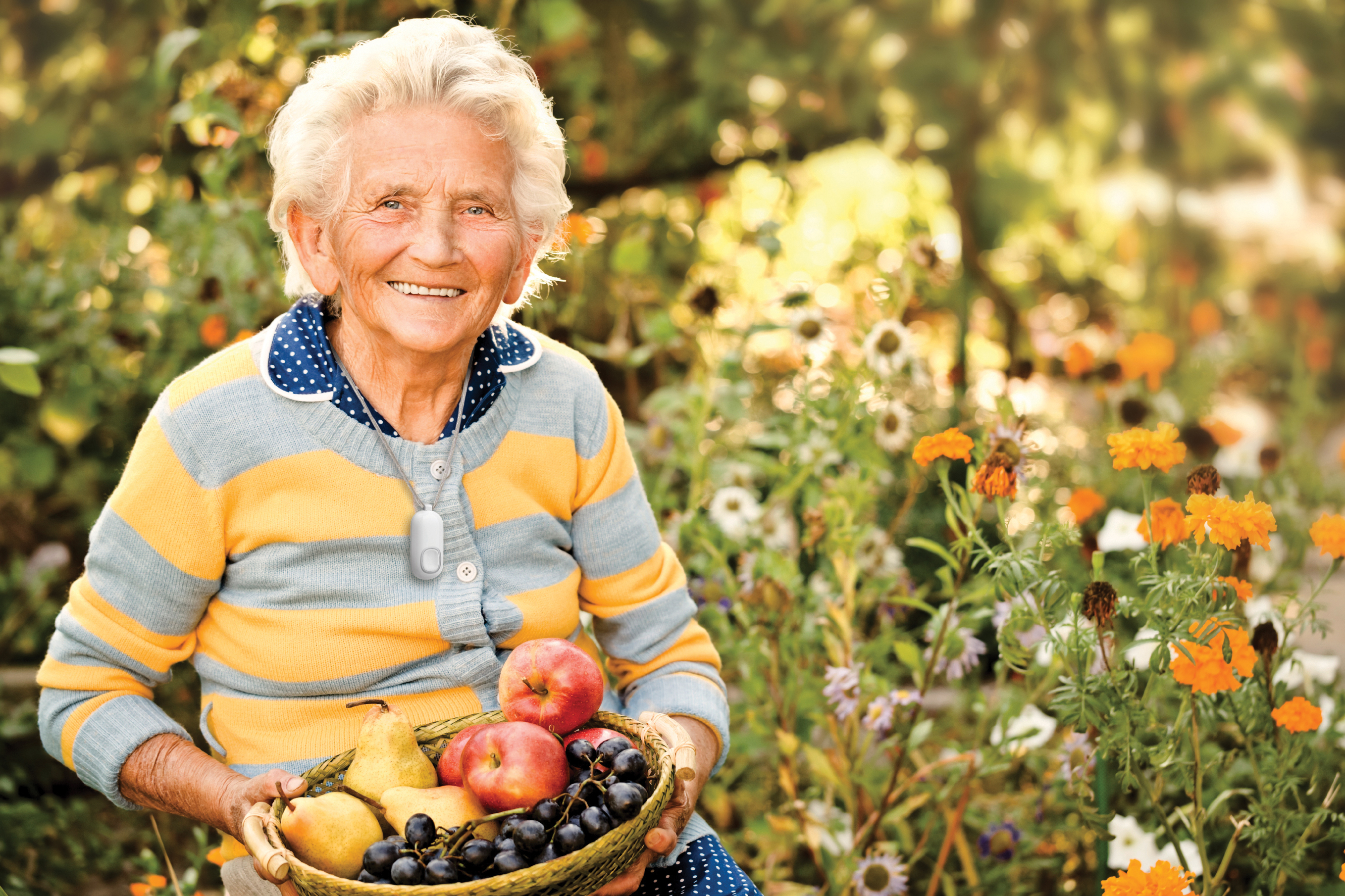 Smiling older woman wearing the Auto Alert fall detection button sitting in a field of flowers holding a basket of fruit.